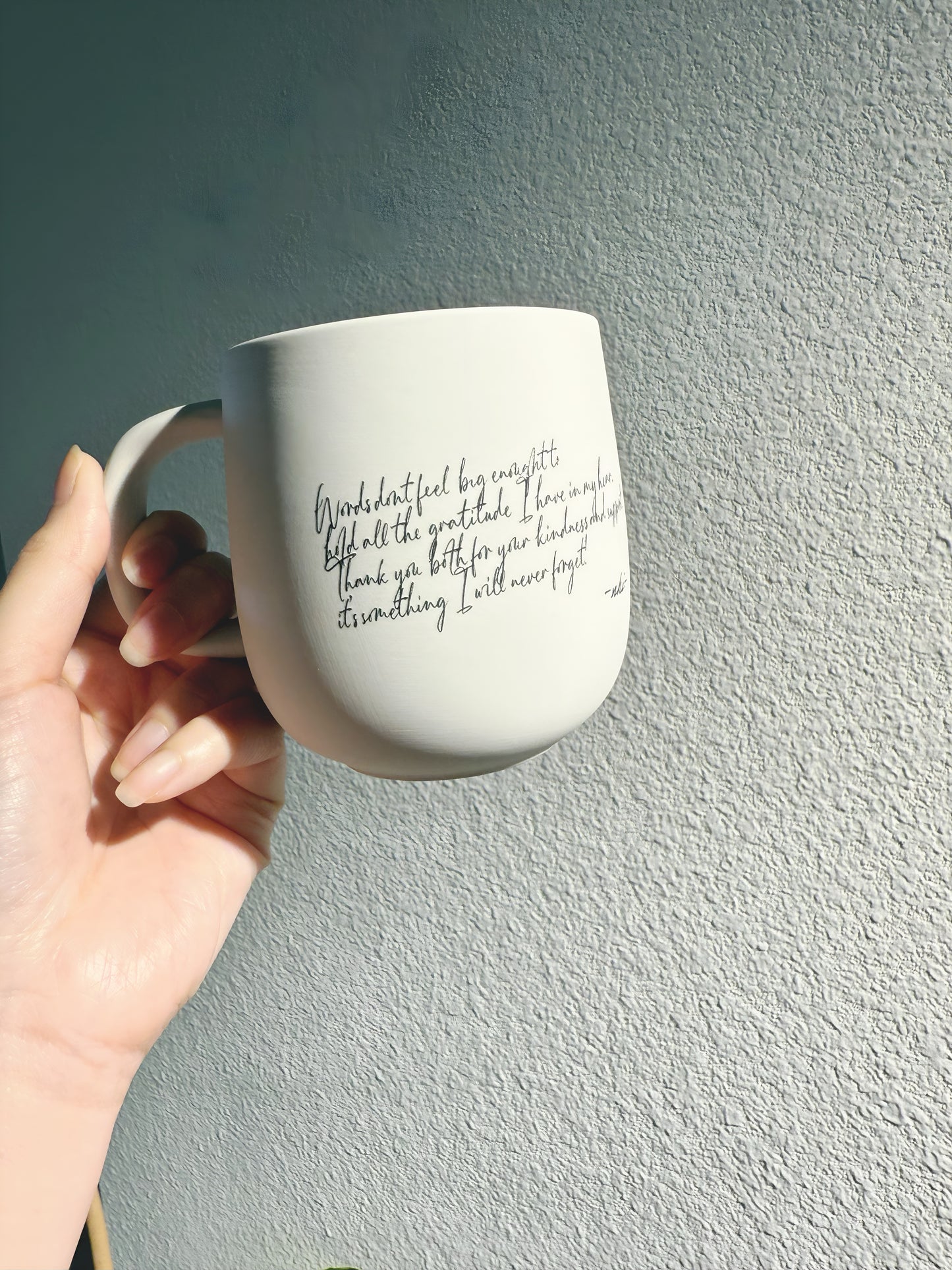 White mug with text held by a hand against a gray wall