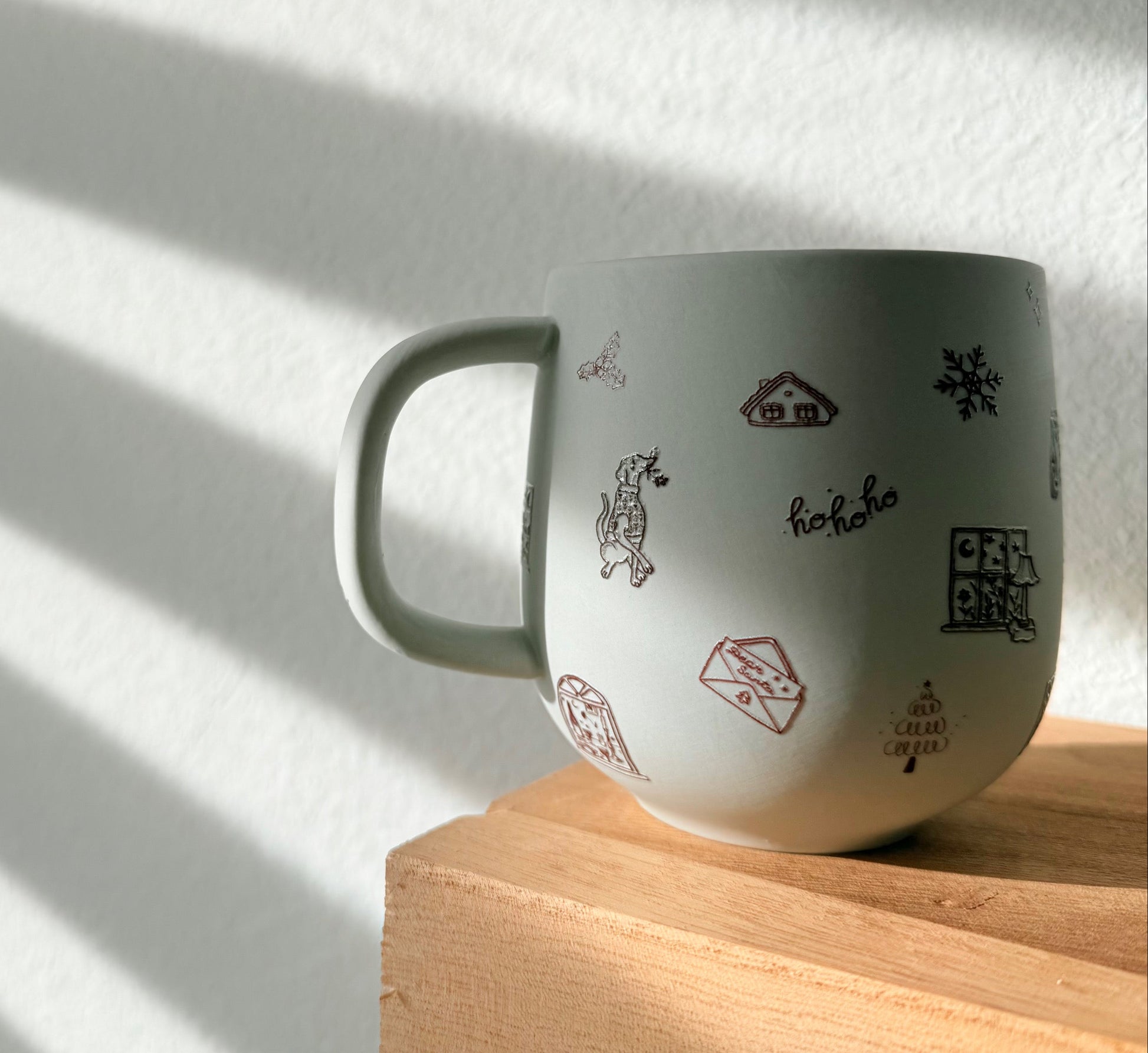 White mug with black and red designs on a wooden surface with sunlight casting shadows.