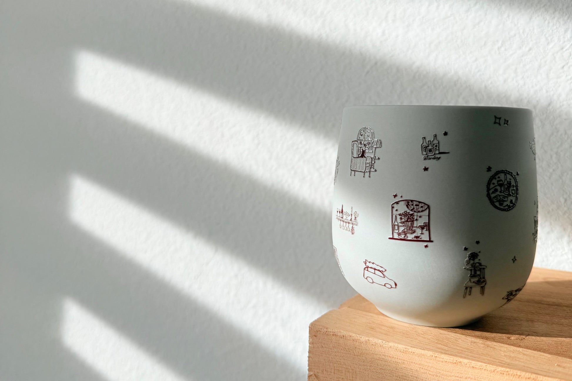 White ceramic cup with black text and symbols on a wooden shelf with sunlight casting shadows on a white wall.