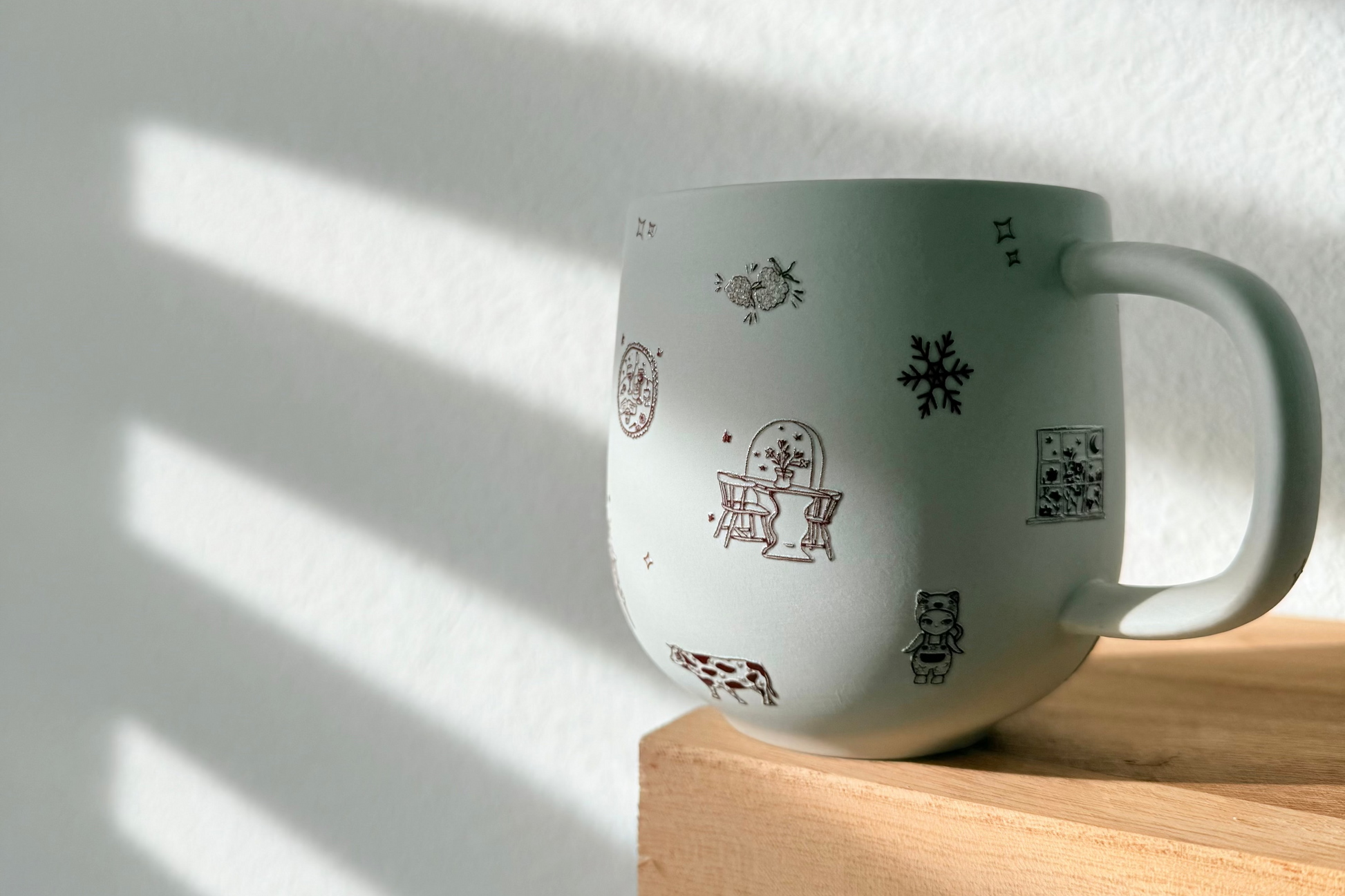 White mug with black and red designs on a wooden shelf with sunlight casting shadows on a white wall.